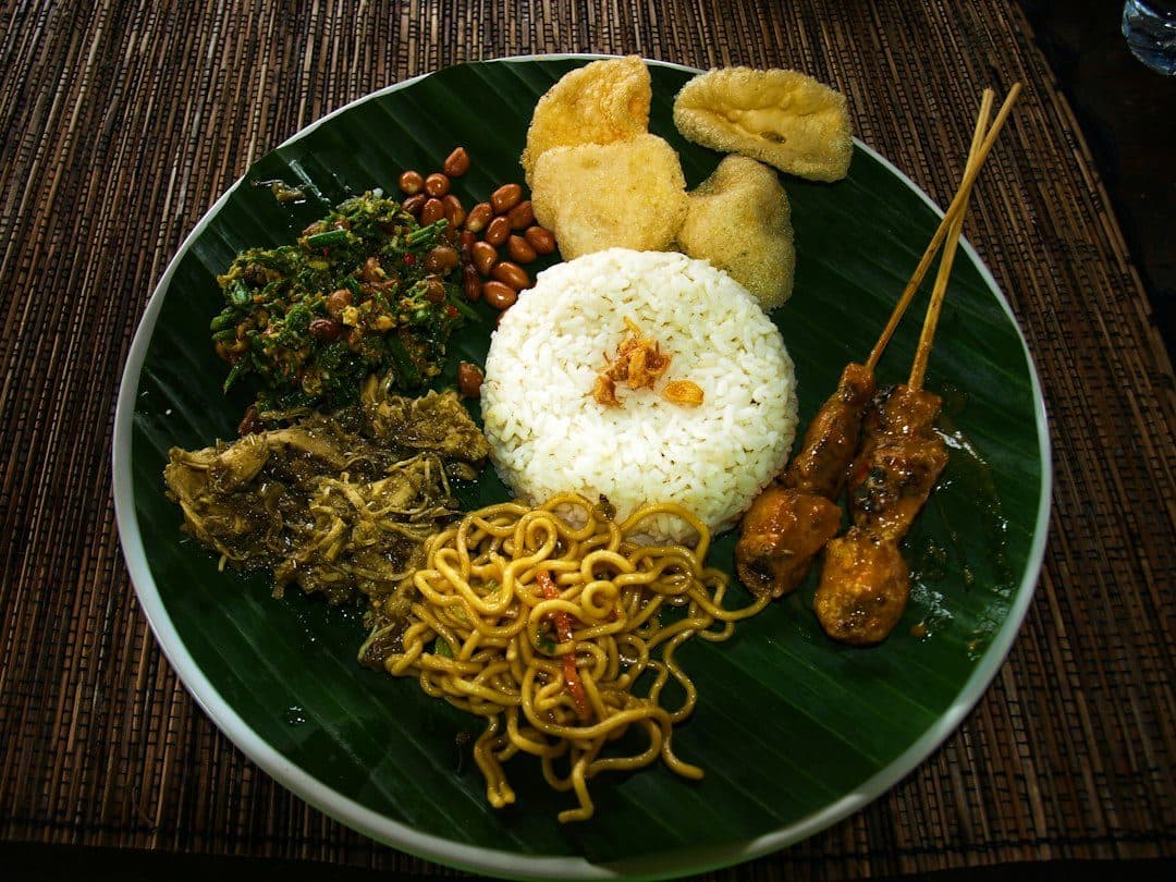 Nasi dengan mi, sate, kerupuk, dan lauk di atas daun pisang