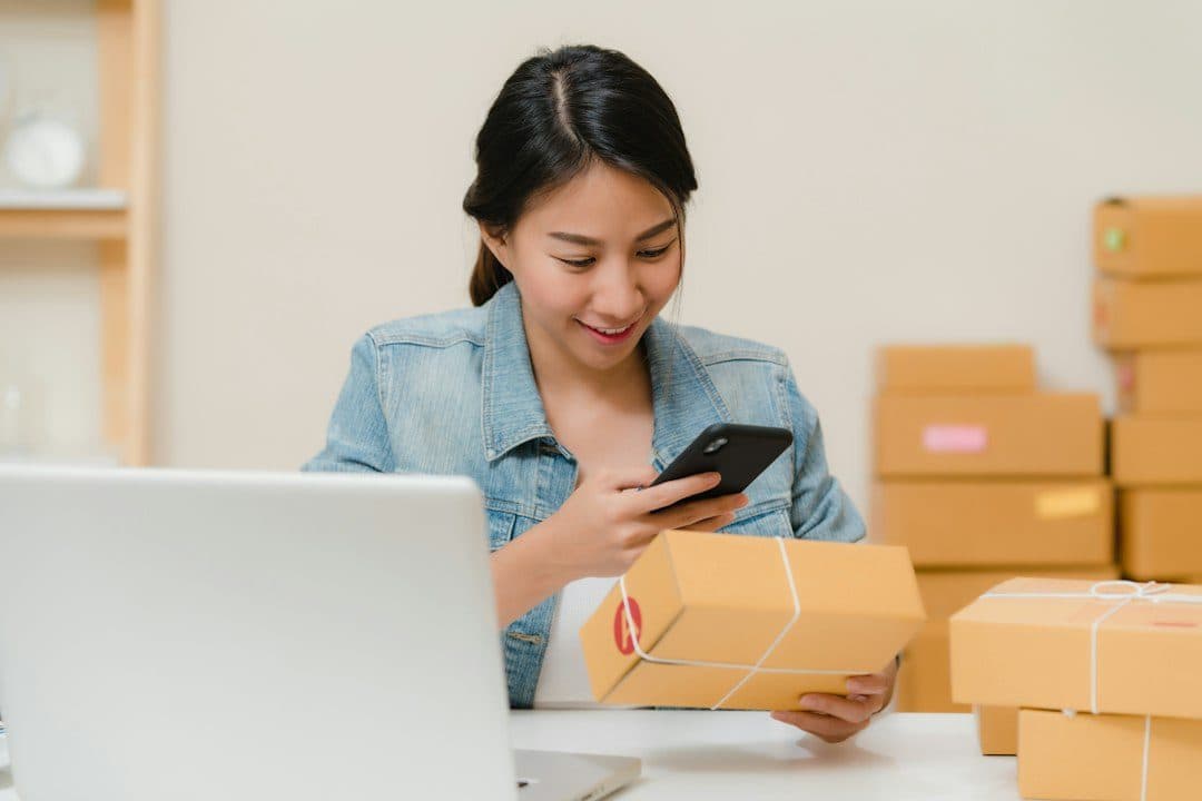 Penjual tersenyum melihat pesanan di HP sambil memegang paket di samping laptop